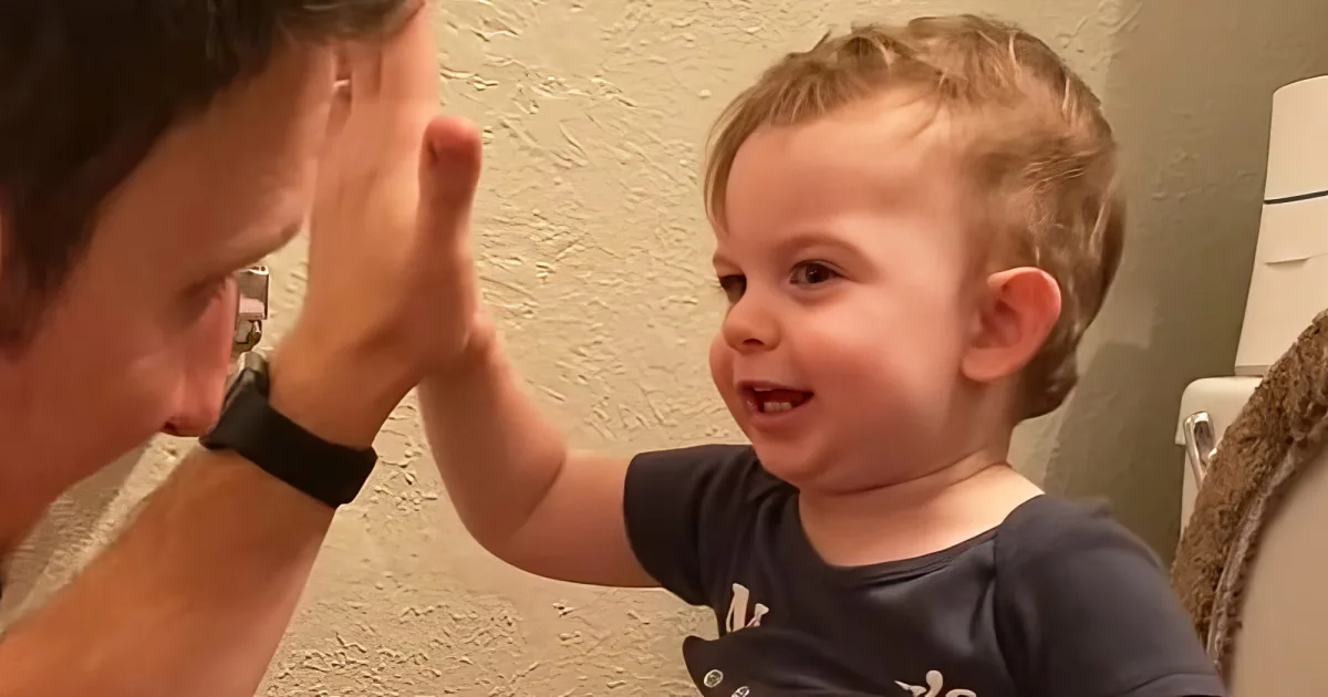 A smiling child in a dark shirt interacts with an adult, both with hands raised, in a warmly lit room.