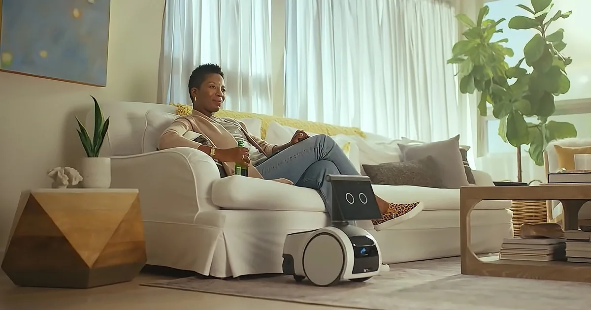A woman sits on a couch, relaxing with a drink, while a small robot sits on the floor beside her.