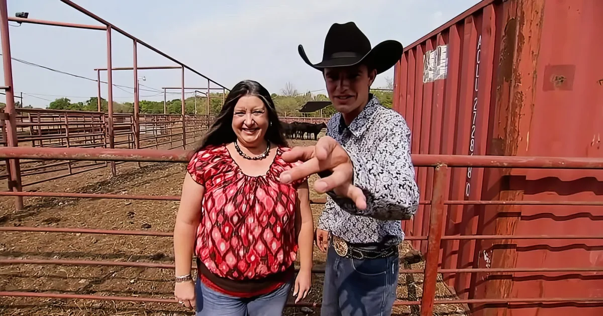 A man in a cowboy hat and woman in a red patterned top stand in a fenced area near a red shipping container.