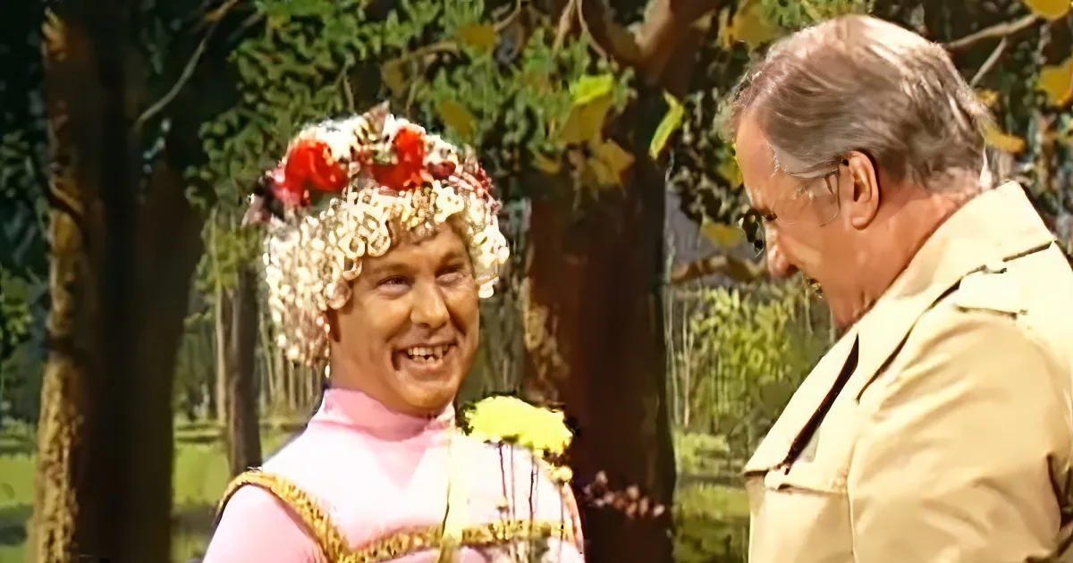 A person in a pink dress with a floral crown and gold belt is interacting with another person in a beige outfit in a...