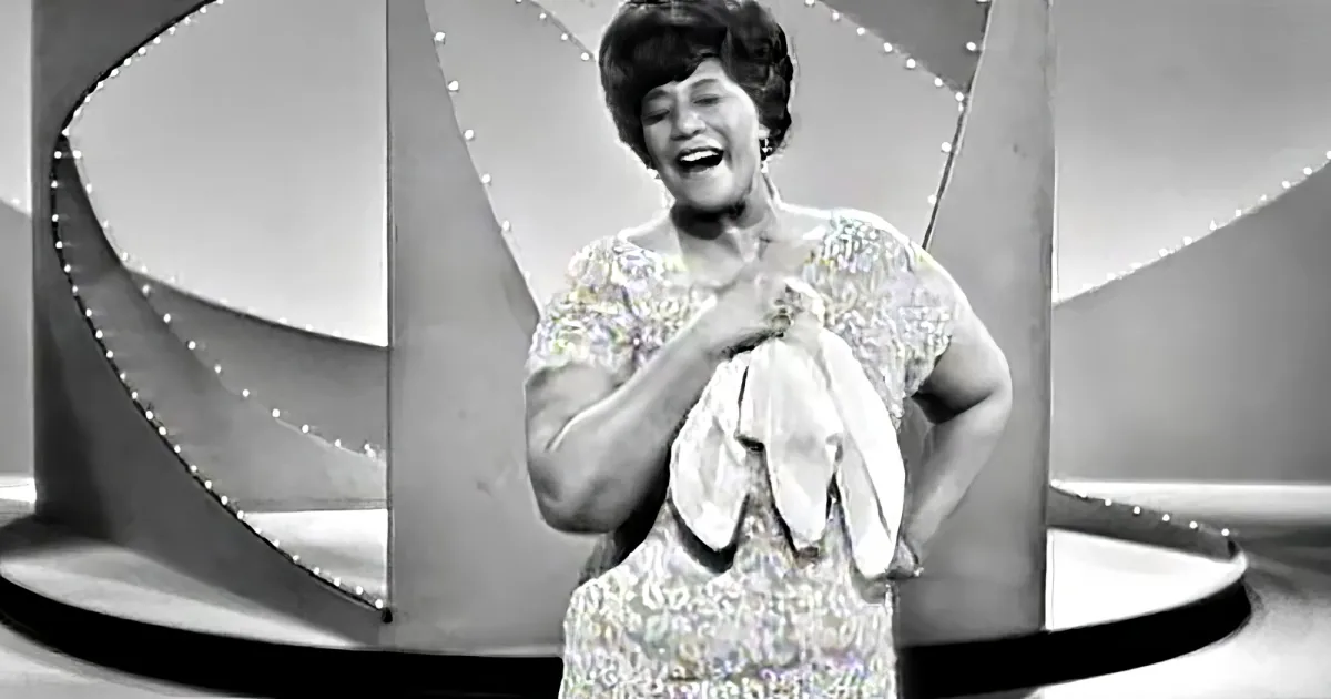 A woman in a sparkling white dress smiles and gestures with her hands on a stage with a decorative backdrop.