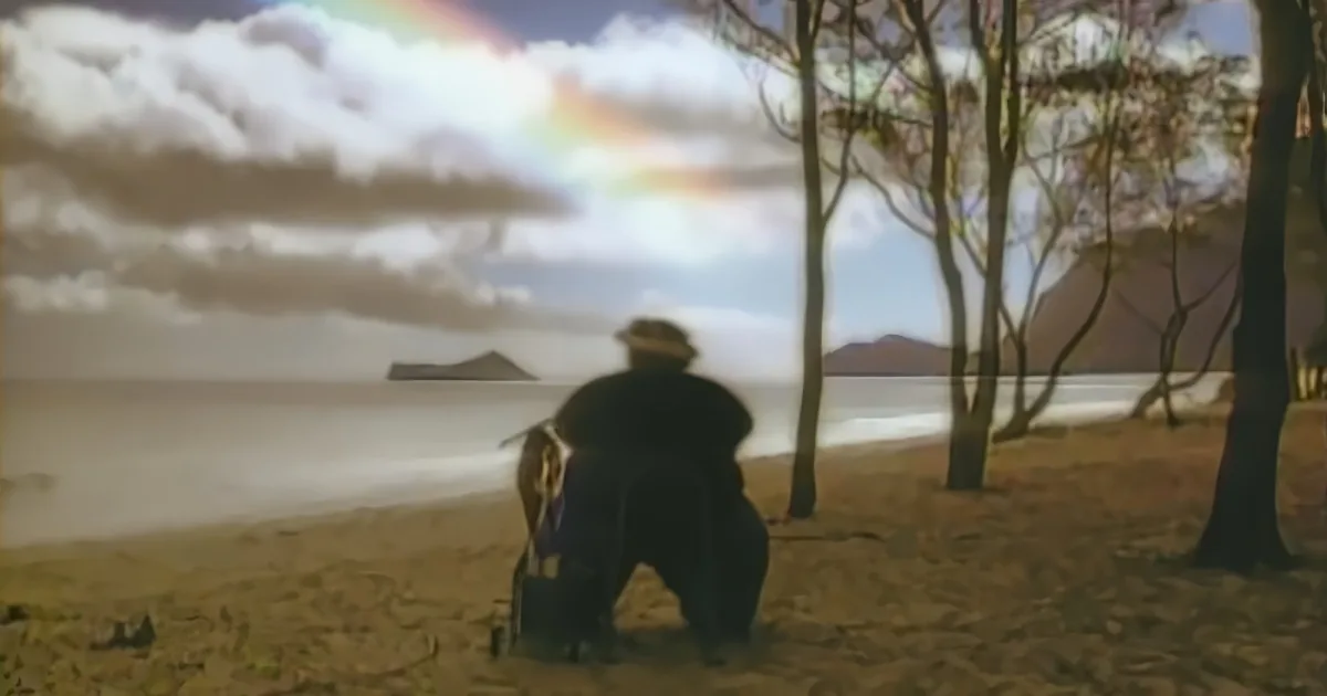 A man in a hat sits on a bench, gazing at a serene beach scene with a rainbow and distant mountains.