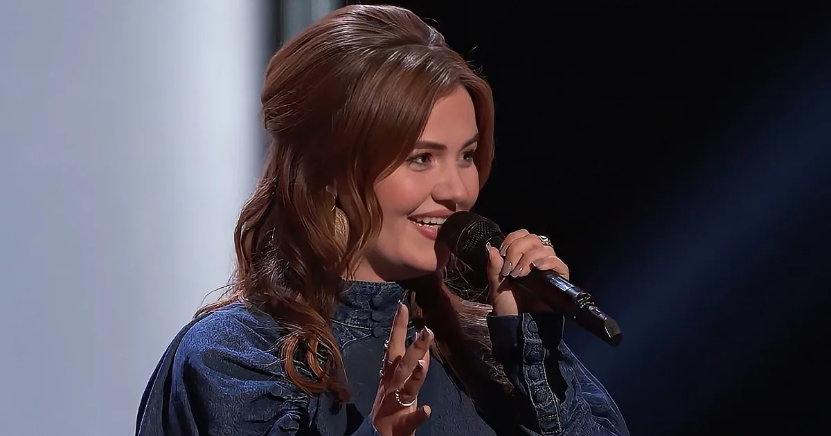 A woman with long brown hair smiles while holding a microphone, wearing a denim jacket.