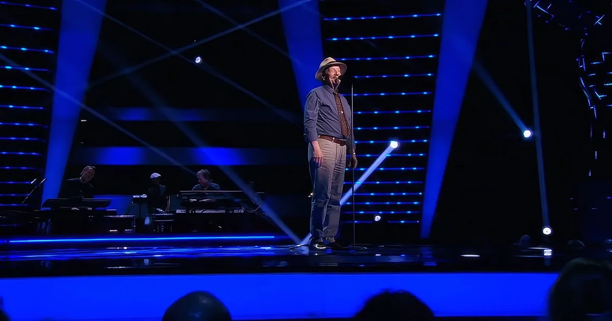 A man in a hat and suit sings on stage with blue lighting and musicians in the background.