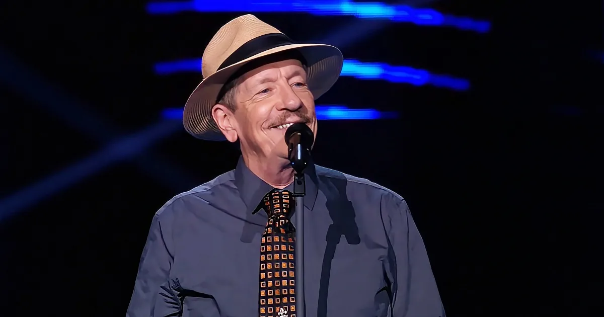 A man wearing a hat and a patterned tie smiles while speaking into a microphone on stage.