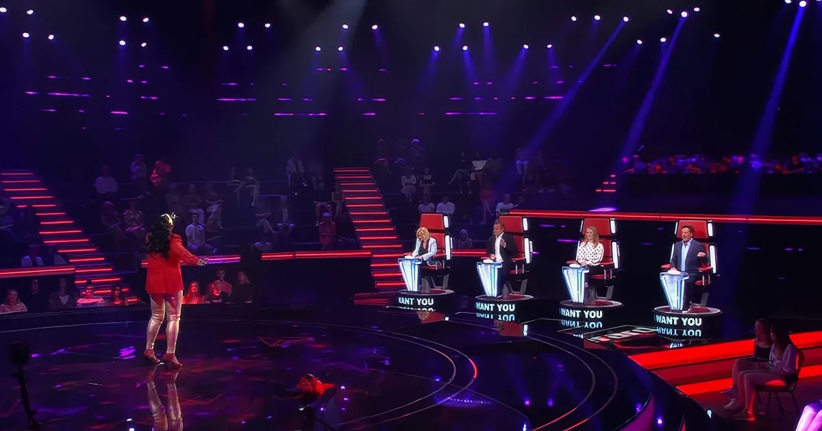 A performer in red dances on stage, with judges seated at podiums and a vibrant audience in the background.