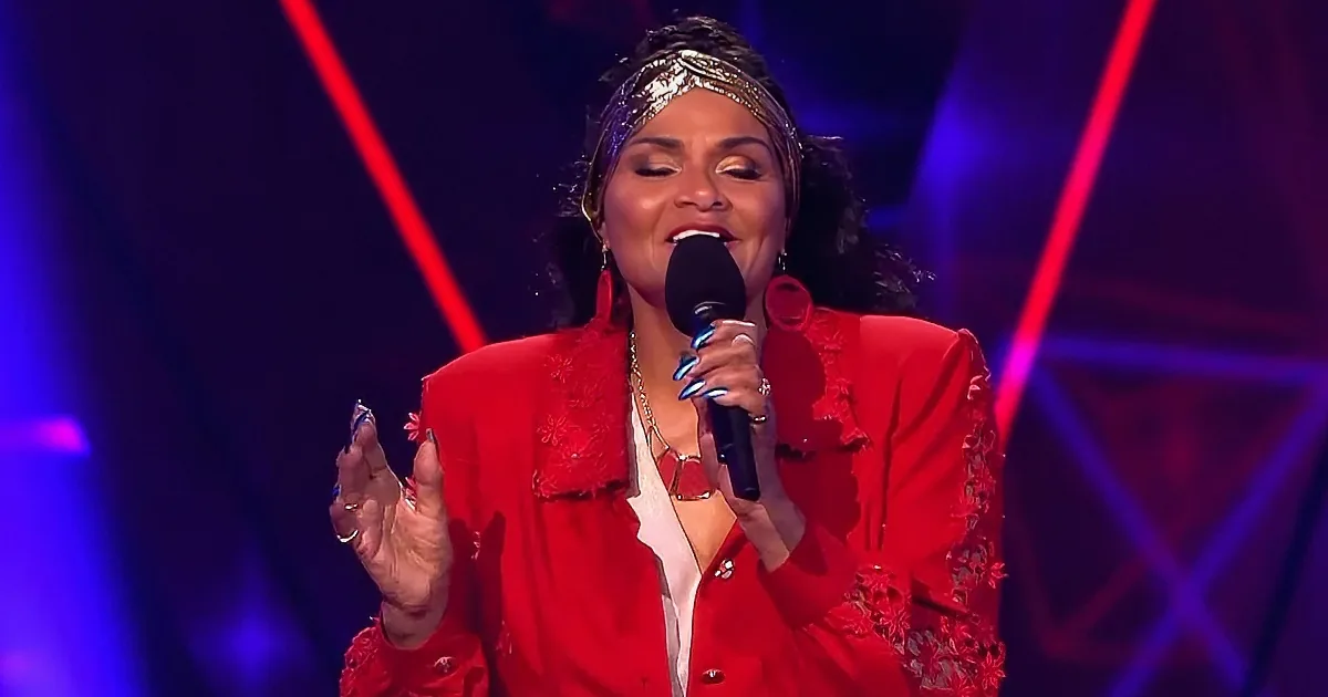 A woman in a red outfit sings passionately on stage with a microphone, surrounded by vibrant stage lights.