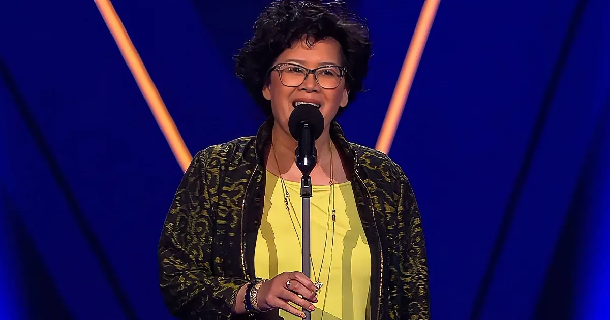 A woman in a patterned jacket and glasses stands on stage, holding a microphone, with a blue background and orange lines.