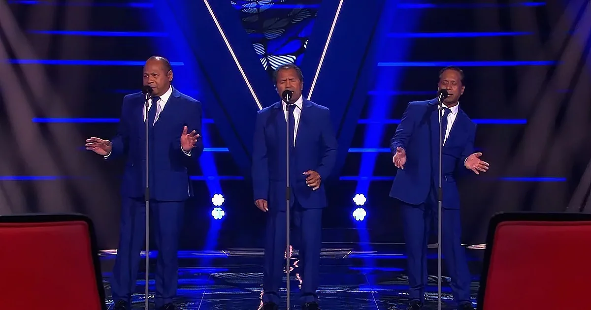 Three men in blue suits sing on stage with microphones, illuminated by blue lights.