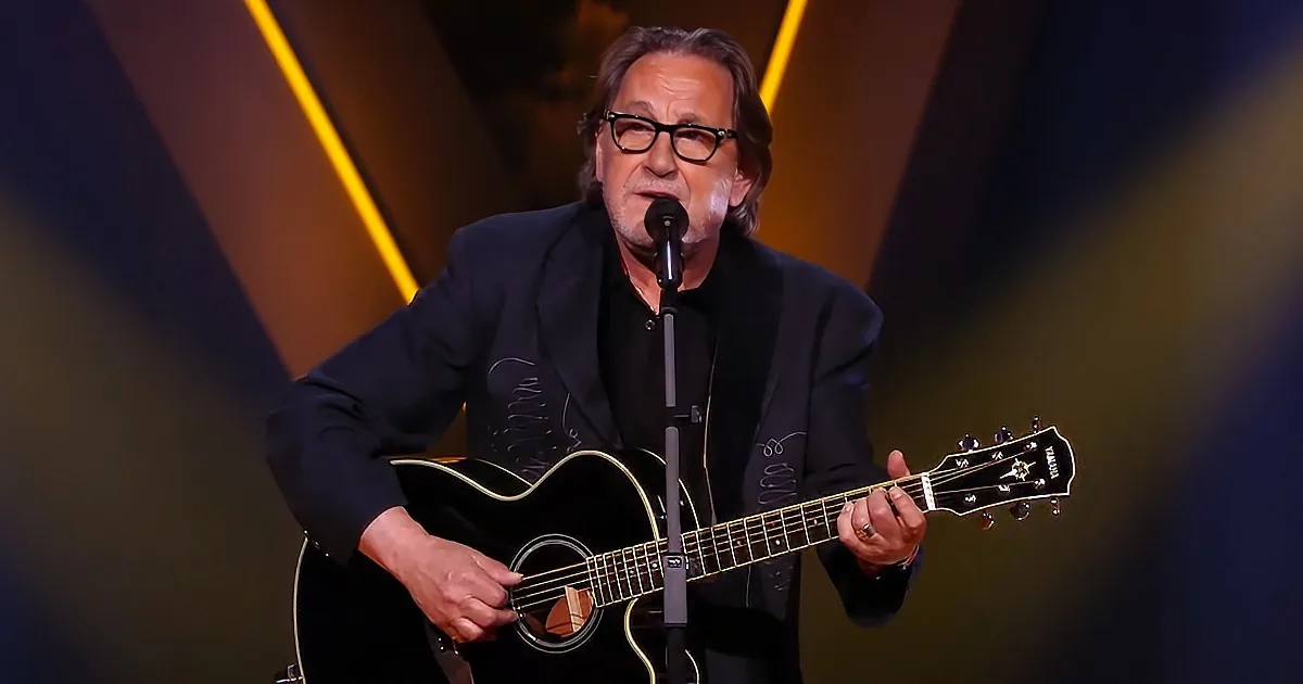 A man in a suit plays an acoustic guitar and sings into a microphone on stage.