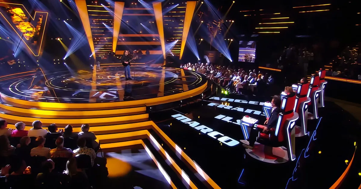 A musician performs on a brightly lit stage with judges' chairs and audience in the background.