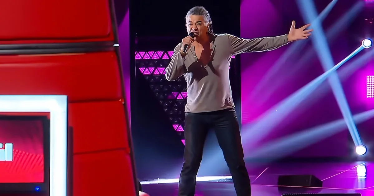 A man in a beige shirt and black pants sings on stage with a microphone, surrounded by vibrant purple and red lighting.