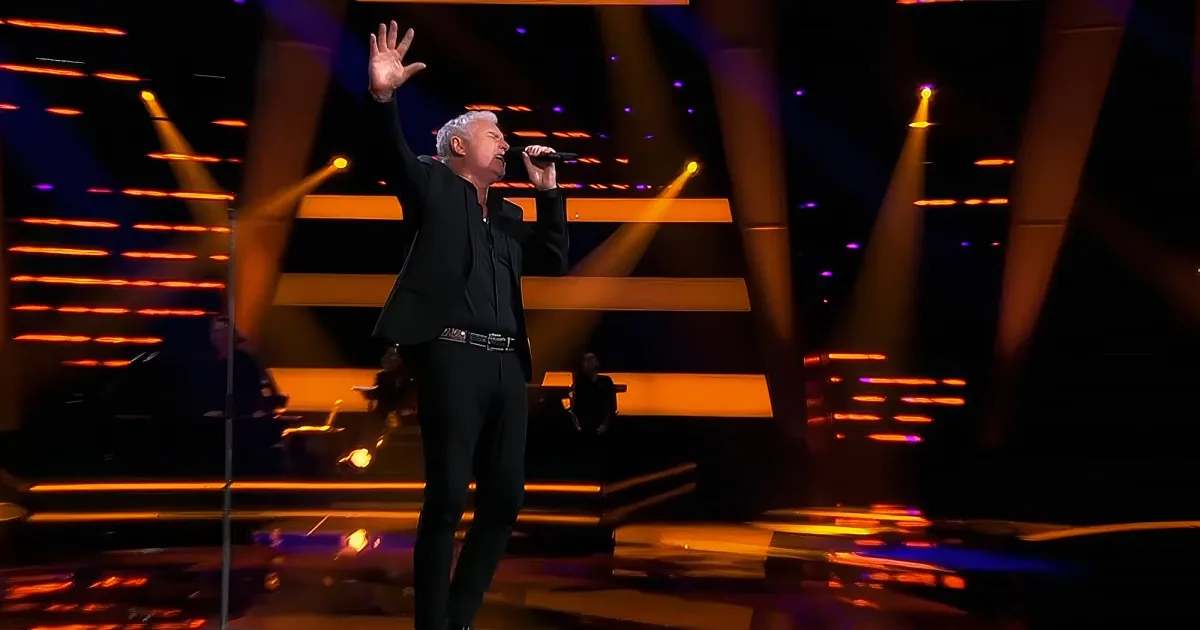 A man in a black suit sings on stage with his hand raised, illuminated by vibrant stage lights.