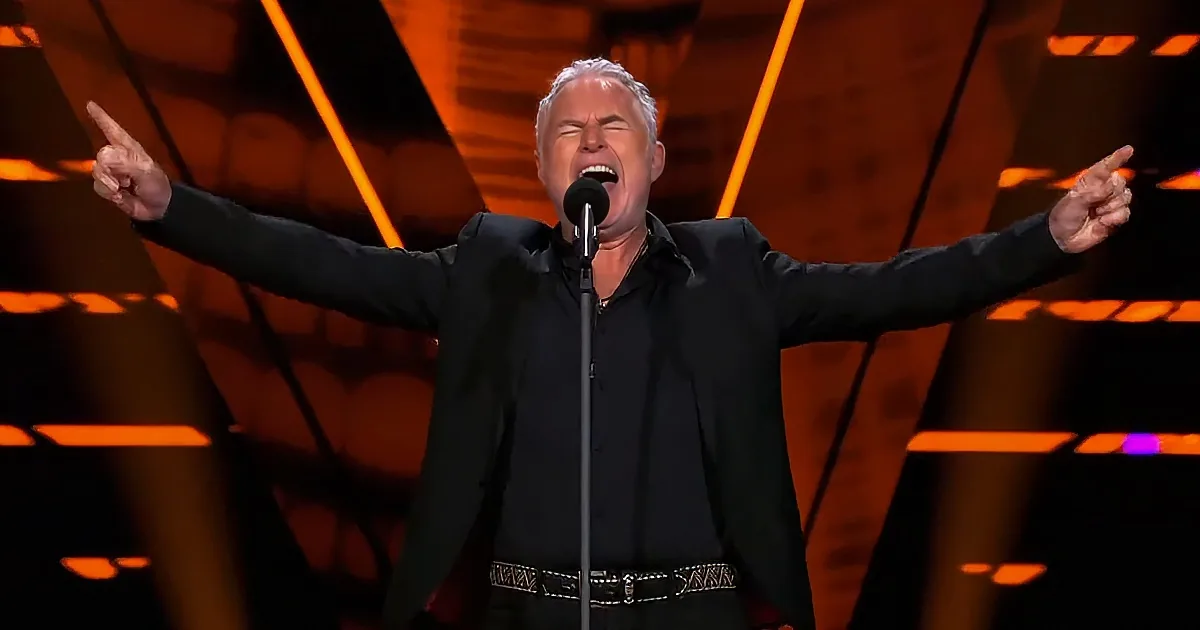 A man in a black suit sings passionately with arms outstretched on stage.
