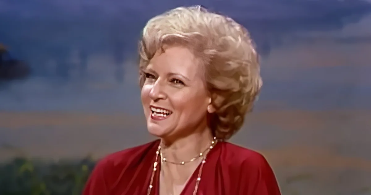 A smiling woman with blonde hair, wearing a red dress and necklace, stands against a blurred background.
