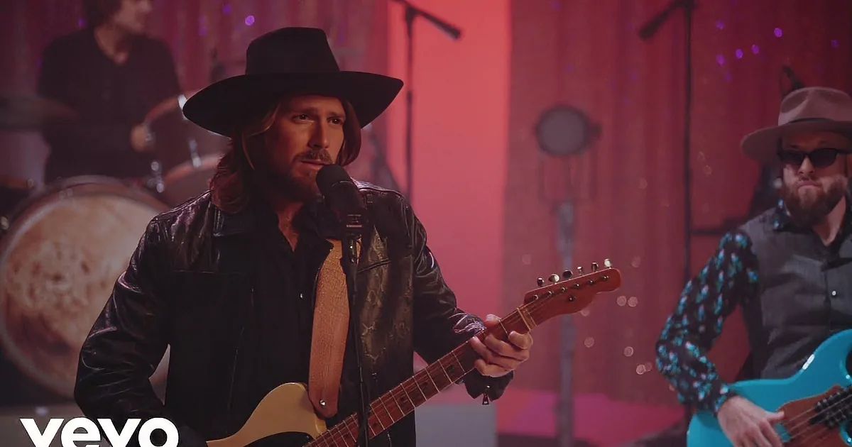 A man in a black hat and leather jacket sings into a microphone while playing a guitar on stage.