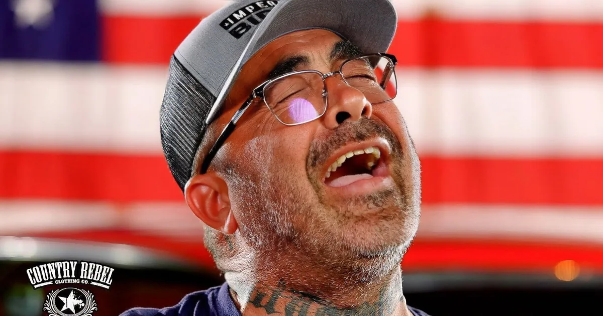 A man with tattoos plays guitar and sings into a microphone in front of an American flag.