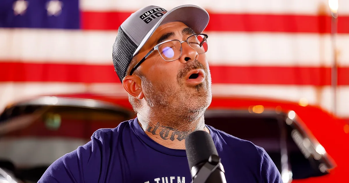 A man wearing a cap and glasses, speaking into a microphone, with an American flag in the background.