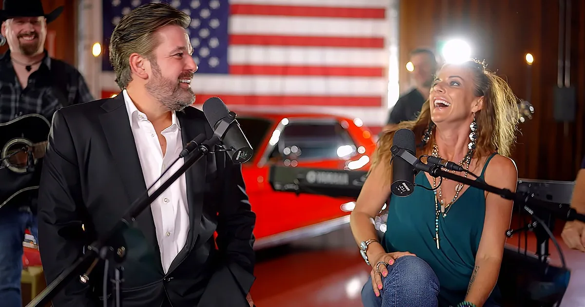 A man and woman sing into microphones, smiling, with an American flag and red car in the background.