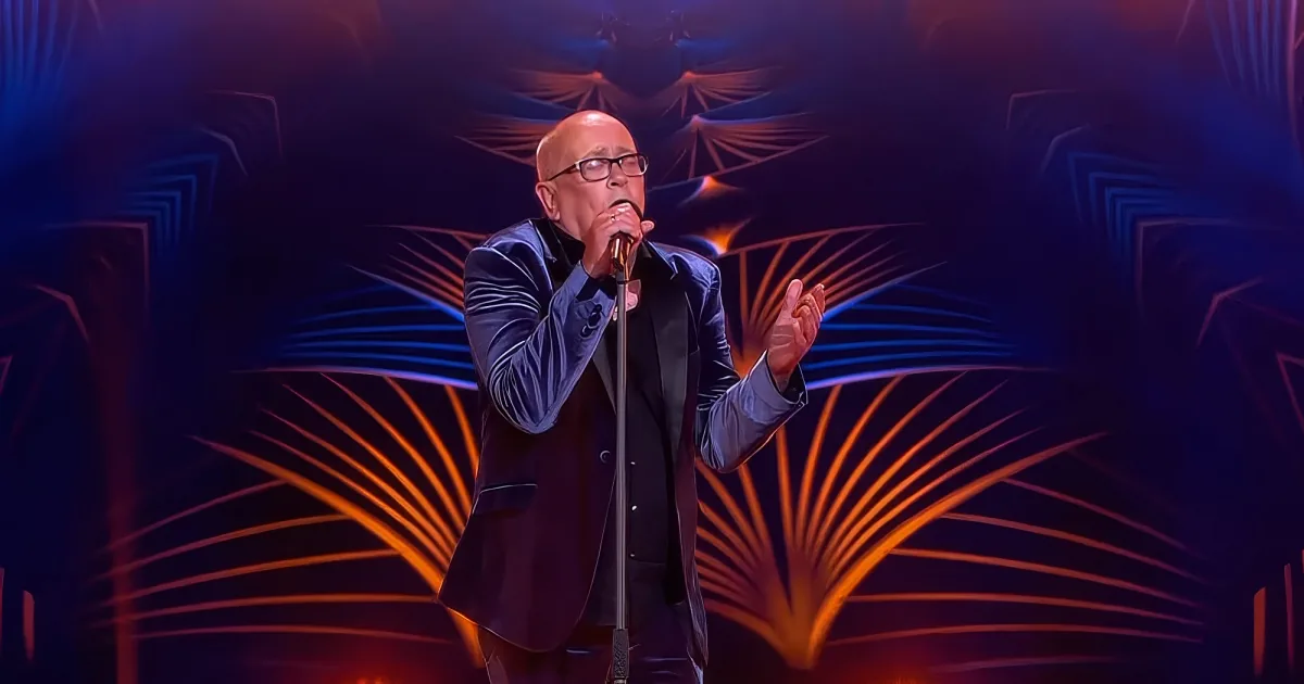 A man in a suit sings into a microphone on stage with a vibrant, abstract background.