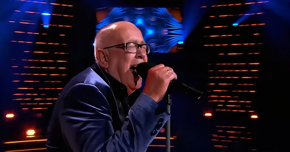 A man in a suit sings passionately into a microphone on stage with vibrant lighting.