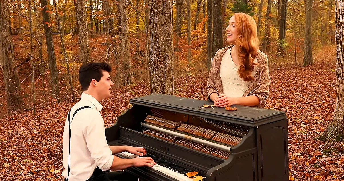 A man plays piano in a forest, while a woman stands nearby, both surrounded by autumn leaves.