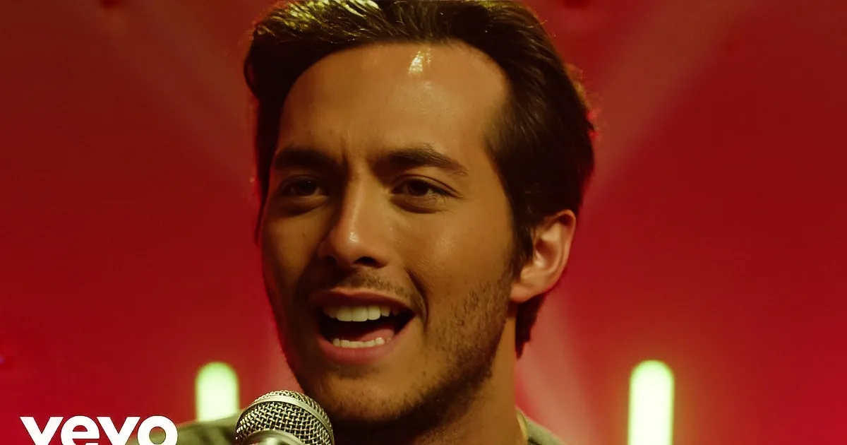 A man in a green shirt sings passionately while playing a guitar on stage under red and green lights.
