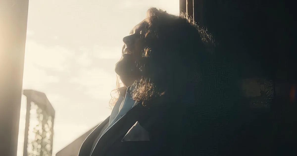 A person with curly hair and a beard looks up, bathed in sunlight, wearing a blue jacket.