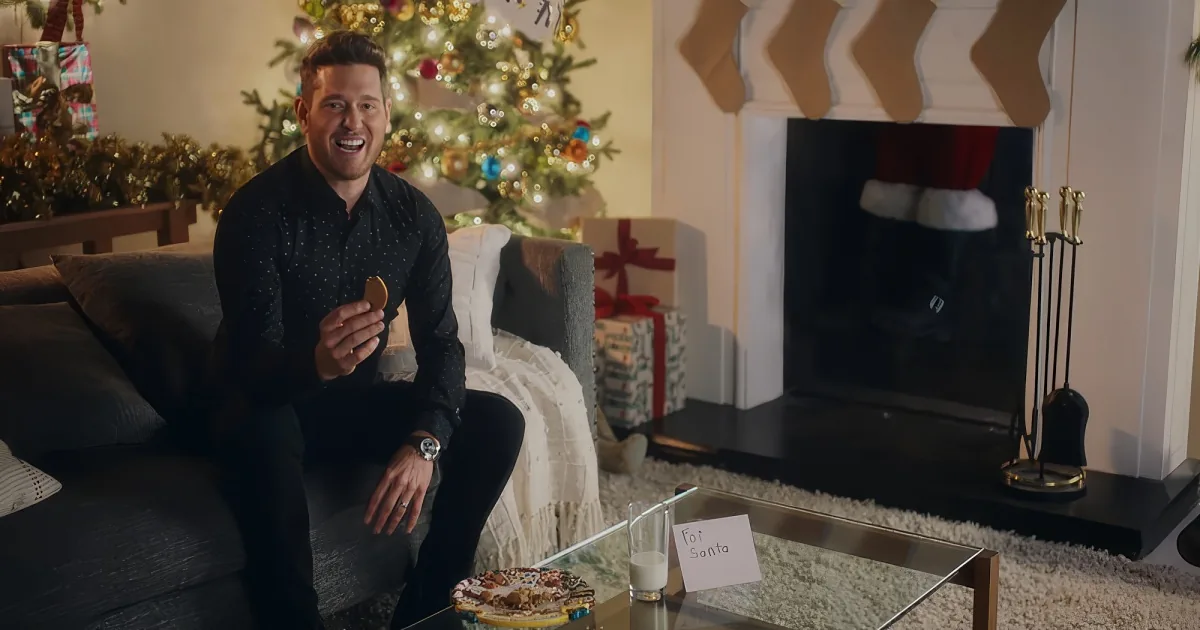 A man sits on a couch, smiling, holding a cookie, with a decorated Christmas tree and stockings in the background.