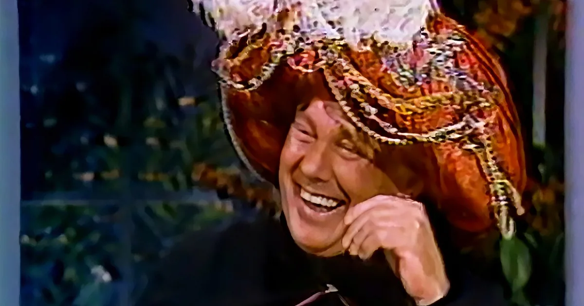 A person wearing a colorful, ornate headdress laughs heartily, with a blurred background of foliage.