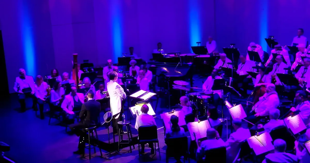 An orchestra with musicians in white attire performing on a stage with blue lighting.