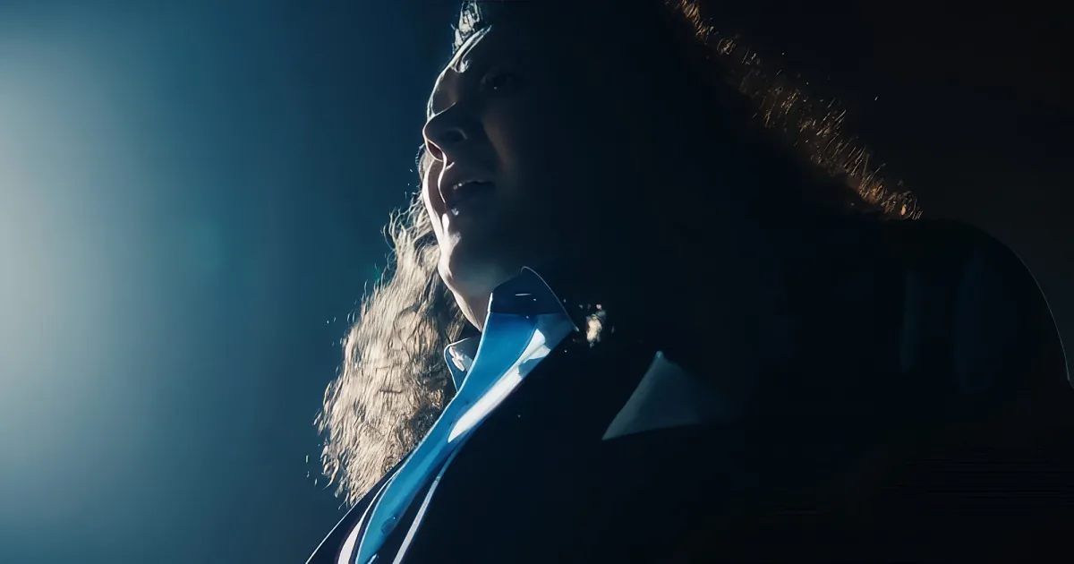 A person with long hair and a blue shirt is shown in a dimly lit environment, possibly singing or speaking.
