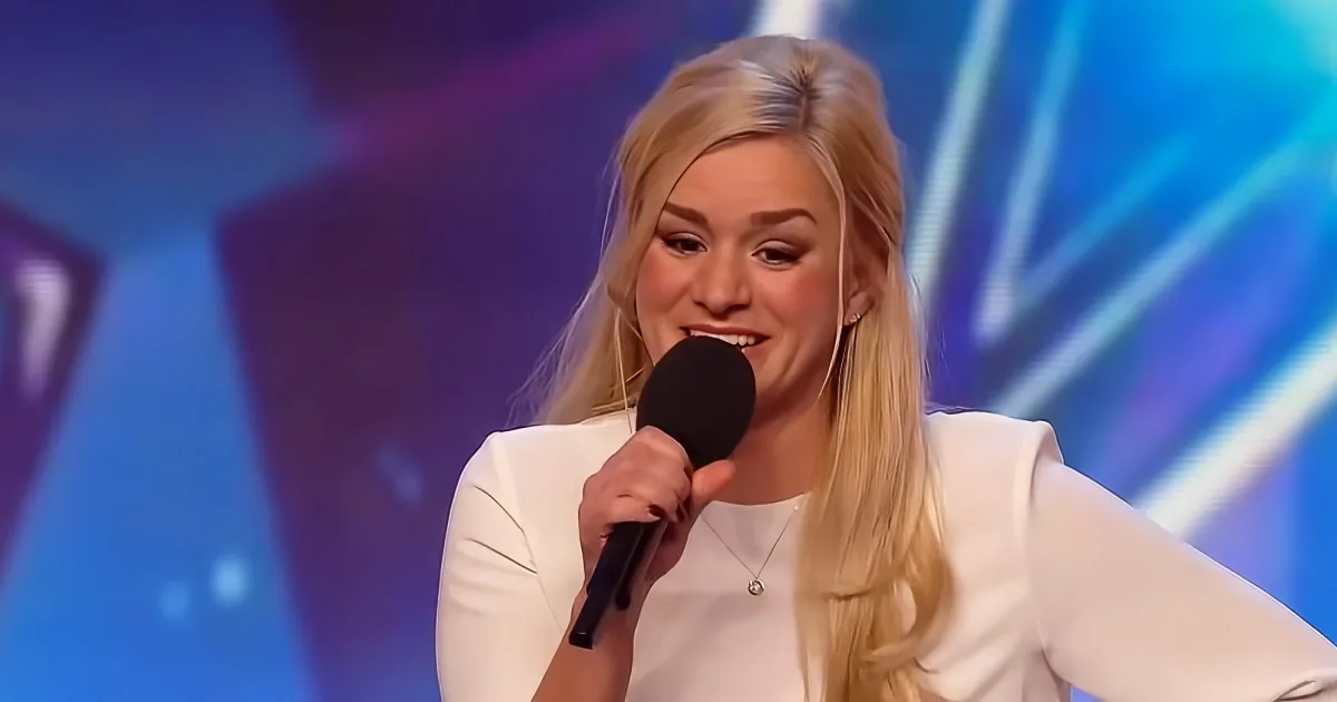 A woman with blonde hair in a white outfit speaks into a microphone against a colorful stage backdrop.