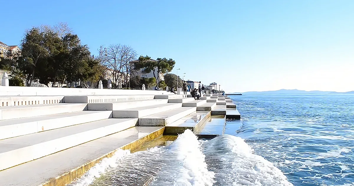 Water flows down steps into the sea, with people and trees in the background under a clear sky.