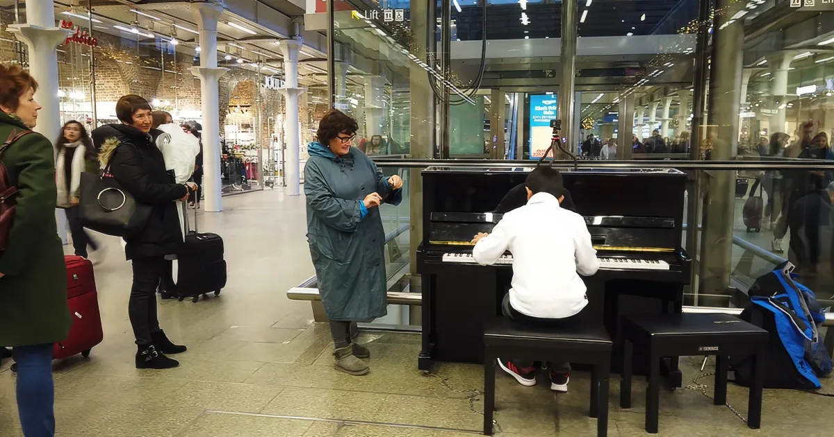 A man in a white hoodie plays a piano in a busy train station, surrounded by onlookers.