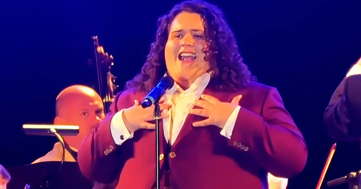 A person with curly hair and a red blazer sings passionately into a microphone, surrounded by musicians.