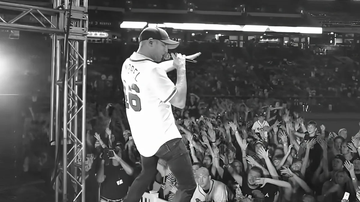A performer in a baseball jersey sings into a microphone on stage, surrounded by an enthusiastic crowd.