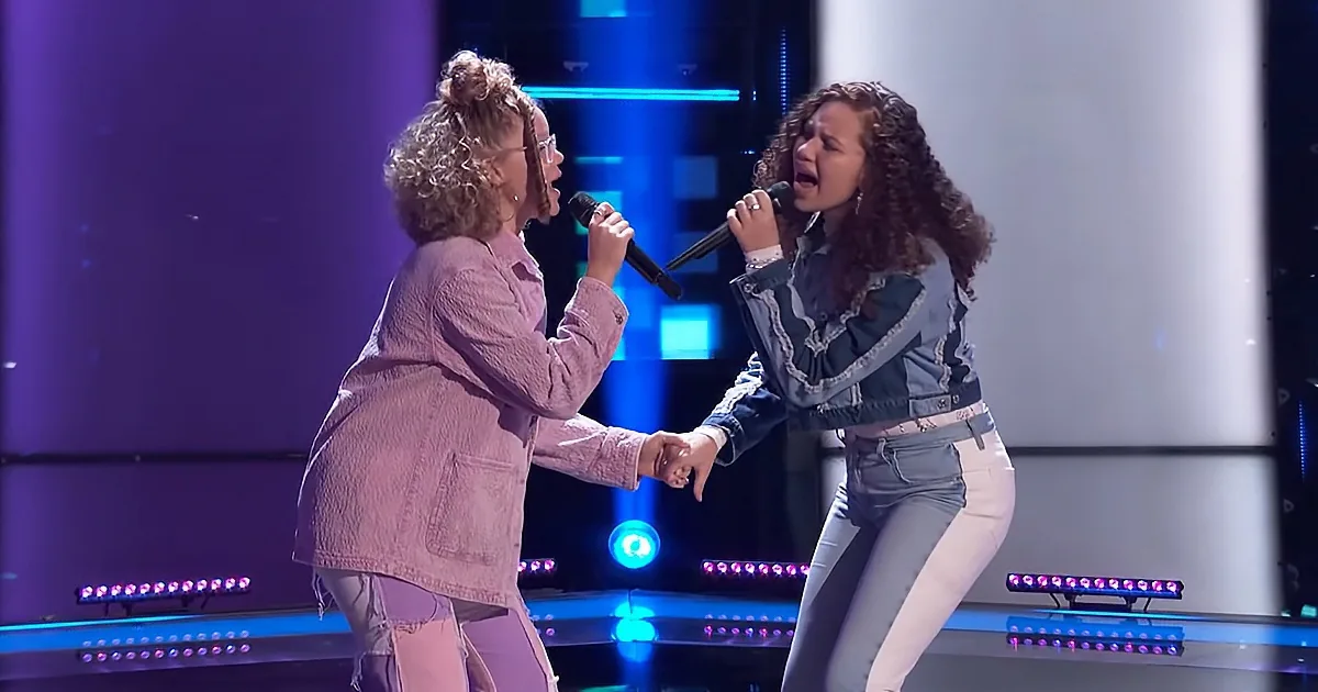 Two women sing passionately on stage, holding hands, wearing casual outfits, with colorful stage lights behind them.