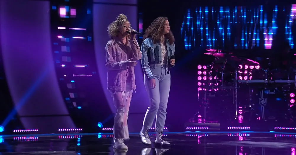 Two women sing on stage with microphones, illuminated by vibrant purple and blue lights.
