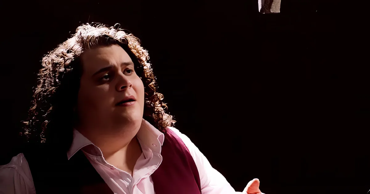 A man with curly hair in a pink shirt and vest is looking up with a surprised expression.