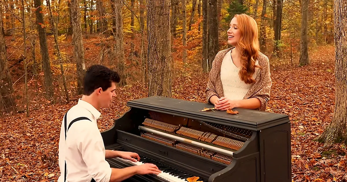 A man plays piano in a forest, a woman watches, surrounded by autumn leaves.