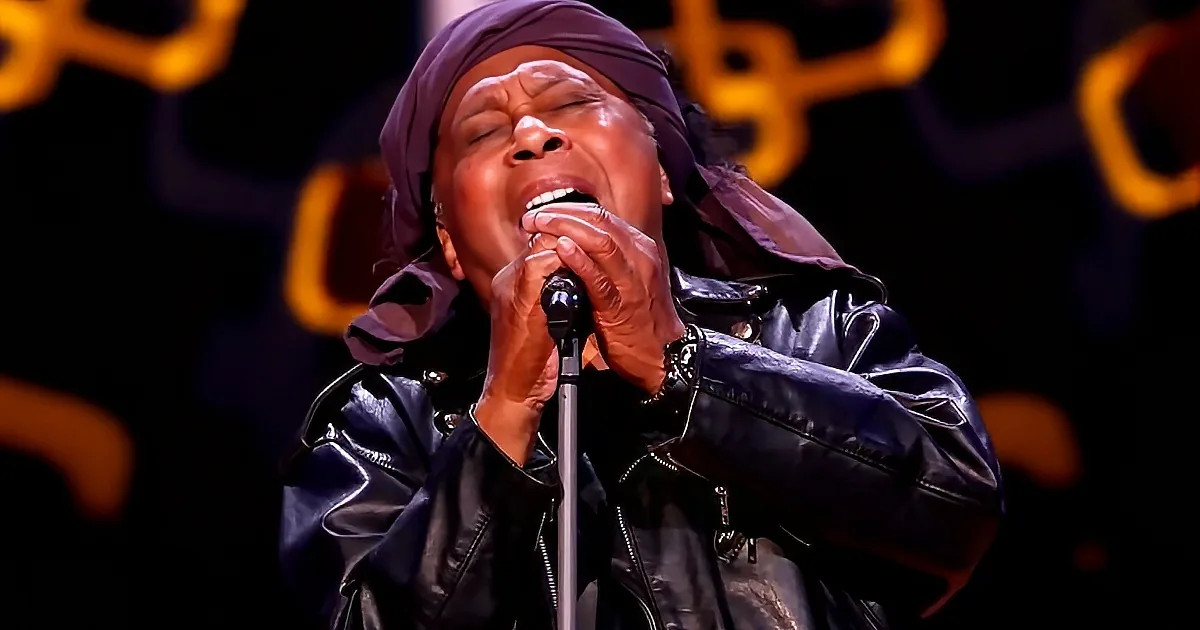 A person in a leather jacket sings passionately into a microphone, wearing a headscarf.