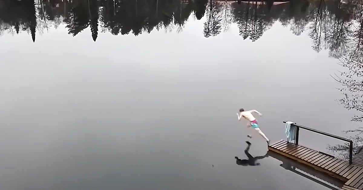 A shirtless person in swim trunks runs off a wooden dock into a calm lake.