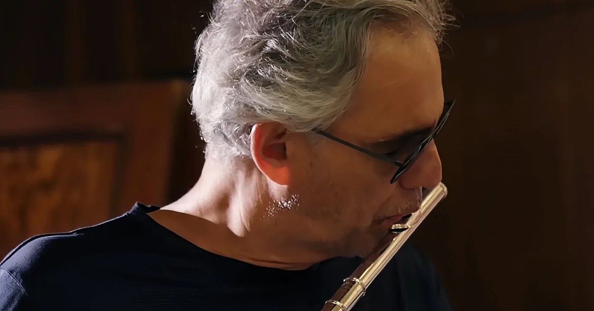 An older man with gray hair and glasses is playing a flute, wearing a dark shirt.
