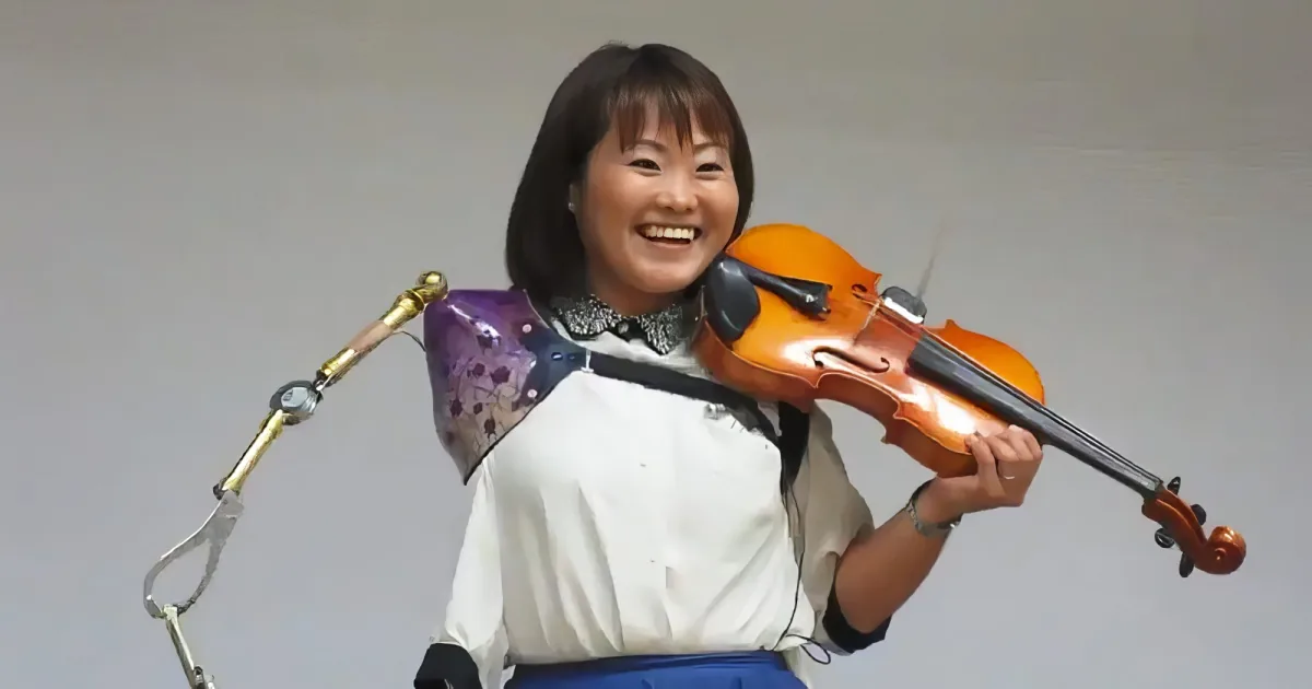 A smiling woman holds a violin, wearing a white shirt and purple floral sleeve, with a gray background.
