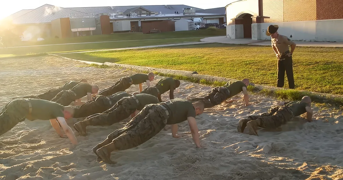 A group of soldiers in green uniforms is doing push-ups on sand under a sunny sky, with a uniformed instructor observing.