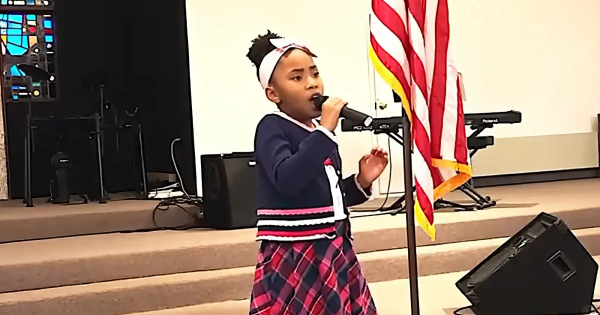 A young girl sings into a microphone on stage, wearing a plaid skirt and a white headband, with an American flag in the...