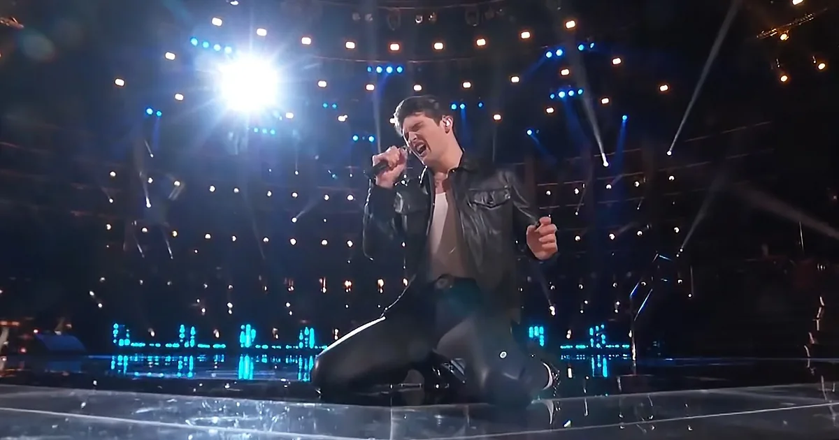 A person in a leather jacket kneels on stage, singing passionately under bright stage lights.
