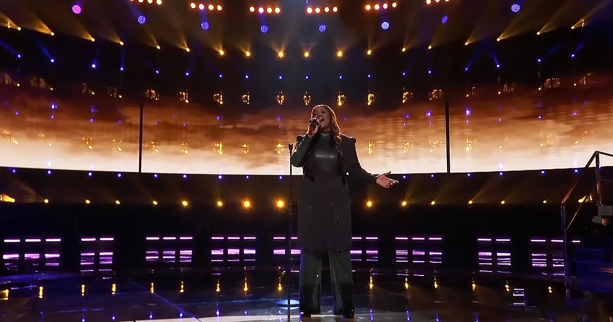 A woman in a black outfit sings on stage with dramatic lighting and a large screen behind her.