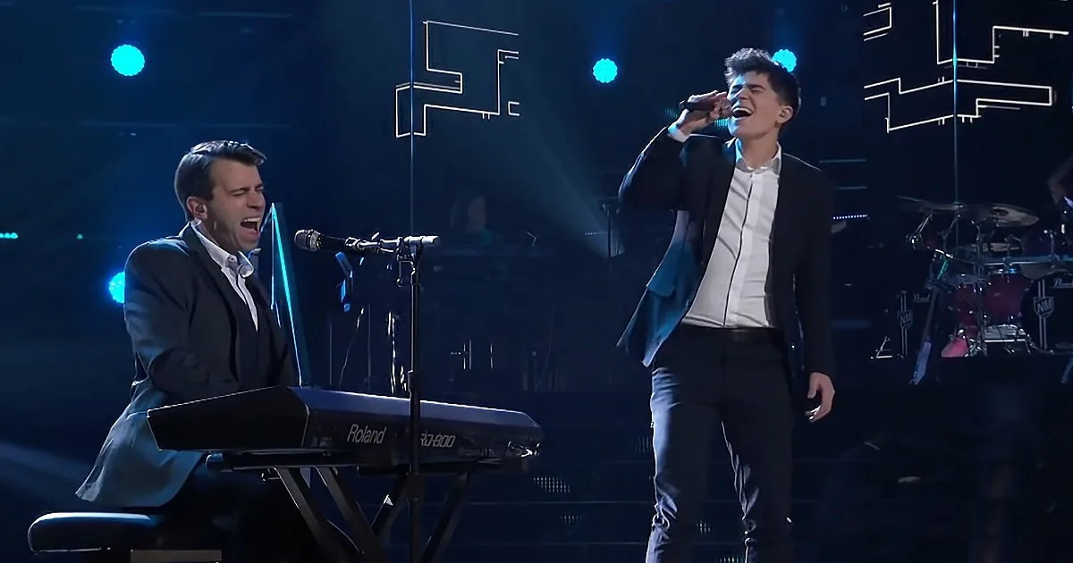 A man sings passionately on stage, accompanied by another playing keyboard, under blue stage lights.